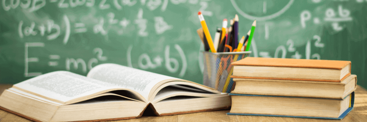 closeup of open textbook, cup of pens and pencils, and stack of books on a school or teacher desk, with a chalkboard with math equations