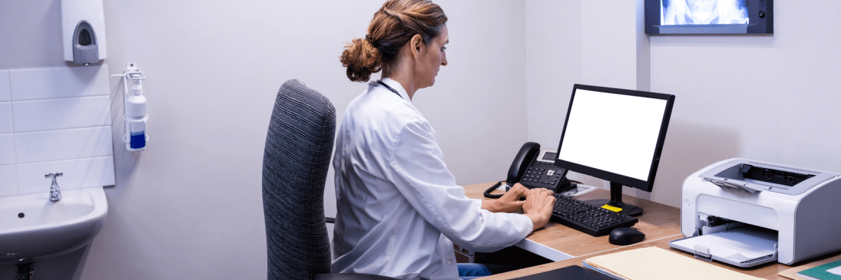 Doctor working on computer with a phone, papers, and a printer on the desk at hospital